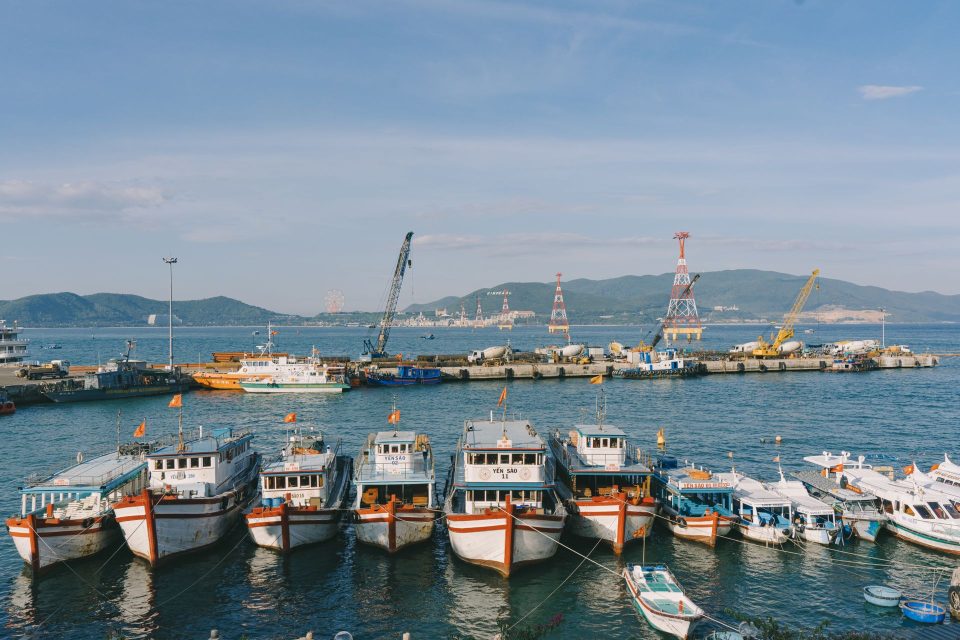 A vibrant harbor scene with fishing boats anchored and cranes in the background under a clear sky.