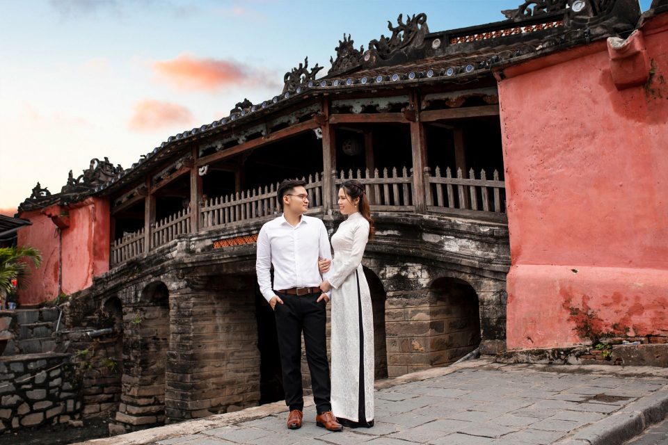 Couple in traditional attire embracing near Hoi An's Japanese Bridge at sunset.