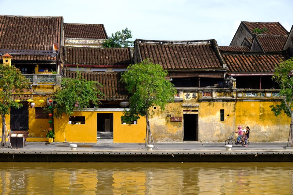 Scenic view of Hội An's historic riverside with vibrant buildings and people biking.