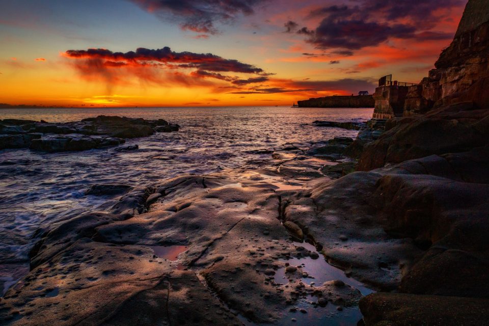 Stunning sunset casting vibrant hues over a rocky shore with dramatic clouds and serene ocean views.