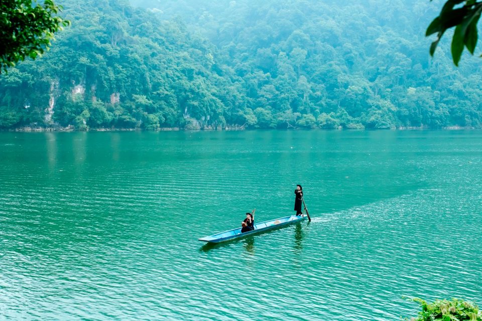Two people in a boat navigate a tranquil, emerald-colored lake surrounded by lush forest.