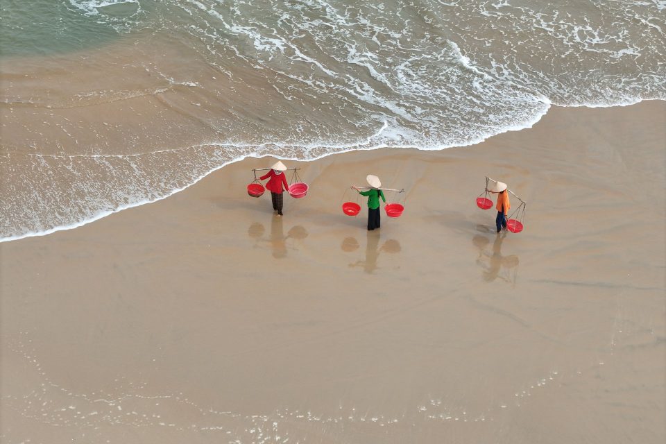 Aerial view of Vietnamese fishermen with conical hats walking on a sandy beach.
