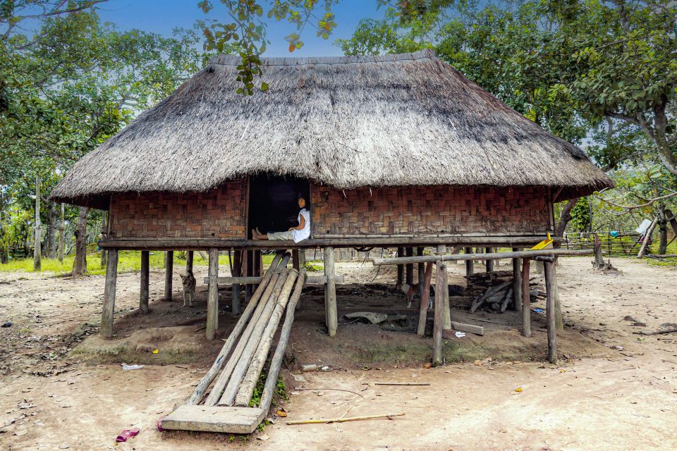 A traditional thatched stilt house in rural Vietnam with a person relaxing in the doorway.