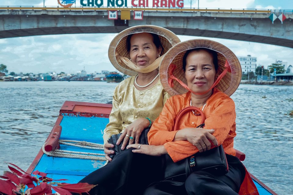 Two women in traditional hats enjoy a sunny river boat ride under the Cai Rang bridge.
